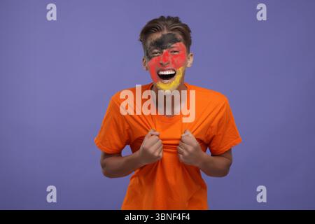 Begeisterter Fan mit der Flagge Deutschlands auf seinem Gesicht vor dunkelviolettem Hintergrund Stockfoto