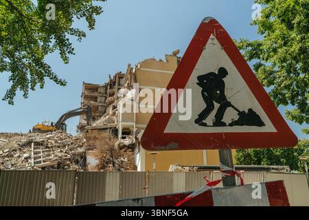 Bauwarnschild vor einem einstürzenden Gebäude und einer aktiven Abbruchstelle mit einem Bagger zur Beseitigung von Schutt. Stockfoto