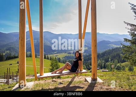 Frau, die sich auf der Schaukel mit Bergblick entspannt. Eine Frau sitzt barfuß auf einer großen Holzschaukel mit Blick auf ein wunderschönes grünes Tal und Berglandschaft Stockfoto