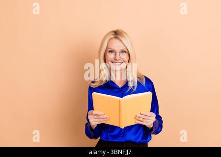 Lächelnde, reife Geschäftsfrau, die ein offenes Buch hält, mit stilvoller blauer Bluse und Brille vor beigefarbenem Hintergrund Stockfoto
