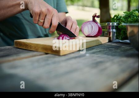 Eine Nahaufnahme der Hände, die eine rote Zwiebel auf einem hölzernen Schneidebrett schneiden, umgeben von frischen Zutaten, im Freien in einer natürlichen Umgebung. Highlights vor dem Essen Stockfoto