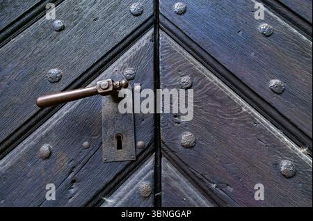 Detaillierte Ansicht einer alten hölzernen Kirchentür mit eisernem Griff, Schlüsselloch und rustikalen Nieten in mittelalterlichem Architekturstil. Stockfoto