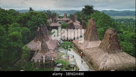 Blick aus der Vogelperspektive auf einzigartige maßgeschneiderte Häuser mit hohen, strohgedeckten Dächern, die ein fesselndes altes Dorf in einer abgelegenen Gegend der Insel Sumba, Indonesien, bilden, mit Einheimischen, die auf einem Pfad spazieren gehen Stockfoto