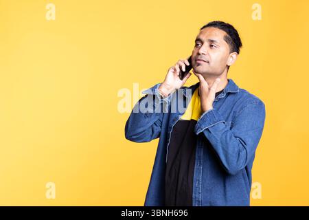 Indische Person lächelt während eines Gesprächs mit seiner Gruppe von Freunden, chelt auf einer Telefonleitung vor orangem Hintergrund. Junger Erwachsener mit einem Männerbrötchen in lässiger Kleidung, Klatsch. Stockfoto