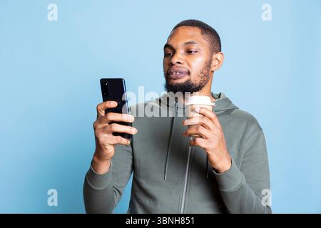 Ein positiver freundlicher Kerl trinkt Kaffee und überprüft sein Smartphone und antwortet fröhlich auf Online-Nachrichten vor blauem Hintergrund. Glücklicher Mann, der gute Nachrichten und Botschaften feiert. Stockfoto
