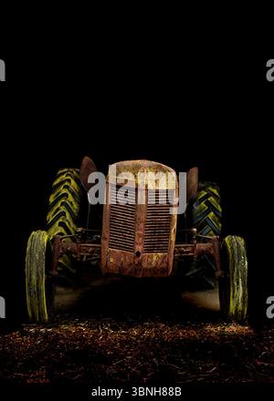 Ein alter, rostiger Traktor wird in dunkler Umgebung gezeigt, dessen Vorderseite zum Betrachter zeigt. Die Reifen des Traktors sind grün und gelb und die Karosserie des Trac Stockfoto