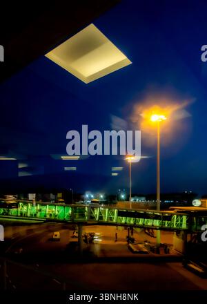Ein Flughafen in der Nacht, mit Leuten, die durch einen überdachten Gang laufen. Der Himmel ist dunkelblau, und es gibt helle Lichter, die die Szene beleuchten. Stockfoto