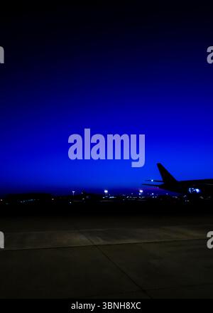 Ein Flugzeug parkt nachts auf dem Rollfeld. Der Himmel ist tief blau und die Lichter des Flughafens sind in der Ferne sichtbar. Stockfoto