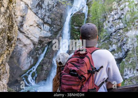 Zwei Touristen mit Rucksäcken, die einen atemberaubenden Savica-Wasserfall beobachten, der in einer wunderschönen natürlichen Umgebung von felsigen Klippen herabstürzt, Slowenien Stockfoto