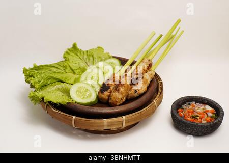 Traditioneller balinesischer Satay lilit, serviert auf Zitronengras-Stäbchen mit frischem Salat, Gurkenscheiben und scharfer Sambal-Matah in einem Steinmörtel. Stockfoto