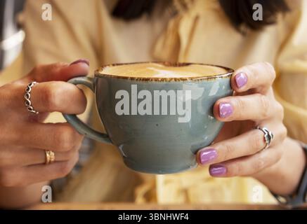 Frau in gelben Kleidern, die mit beiden Händen eine blaue Tasse Cappuccino hält, Nahaufnahme Stockfoto