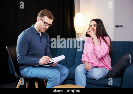 Ein junger Psychologe führt eine Psychotherapiesitzung für eine Frau mit psychischen Problemen durch. Eine Frau sitzt auf dem Sofa und hält ein Glas Wasser hinein Stockfoto