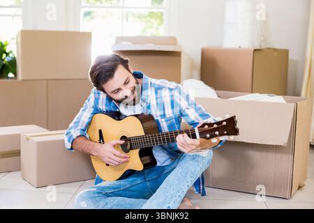Bärtiger Mann, der Akustikgitarre auf dem Boden spielt, mit Umzugskästen, die in Sonnenlicht von den Fenstern eingewickelt werden Stockfoto