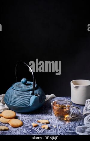 Eine gemütliche Teezeit mit einer blauen Teekanne, einer dampfenden Tasse Tee und köstlichen Keksen auf einem gemusterten Tuch. Ein Milchkännchen verleiht Ihnen einen Hauch von Eleganz Stockfoto