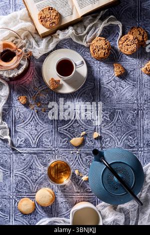 Eine gemütliche Einrichtung mit einer dampfenden Tasse Kaffee, einer Kanne Tee, Schokoladenkeksen, Butterkeksen und einem offenen Buch, alles auf einem Muster Stockfoto