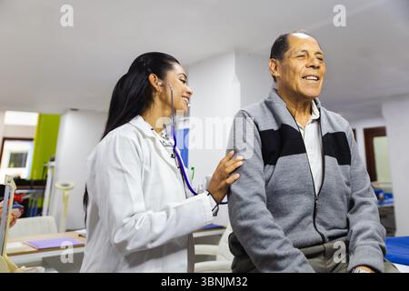 Ein älterer männlicher Patient wird von einer jungen Ärztin in einem lateinamerikanischen Krankenhaus professionell betreut. Der Doktor trägt einen weißen Mantel und Stockfoto