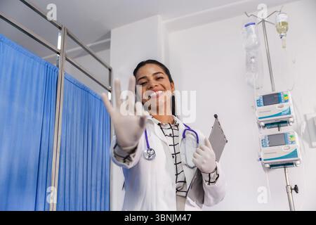 Ärztin in einer medizinischen Umgebung lächelt selbstbewusst, während sie eine OK-Geste zeigt. Sie trägt ein Stethoskop und hält ein Klemmbrett, was Professi symbolisiert Stockfoto
