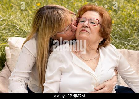 Ein berührender Moment zwischen Mutter und Tochter, die die gemeinsame Zeit in einem sonnigen Park genießen. Die Tochter umarmt ihre Mutter liebevoll und küsst sie Stockfoto