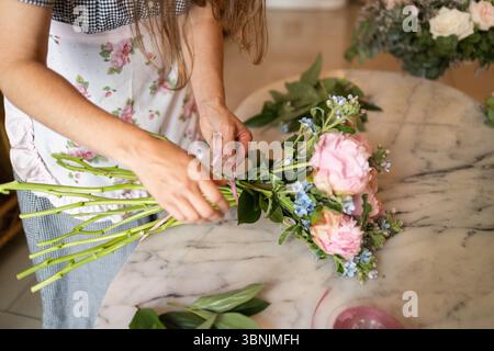 Eine Frau in einem Blumenladen arrangiert geschickt einen wunderschönen Blumenstrauß aus rosa Rosen und blauen Blumen. Ihre Hände binden geschickt die Stiele und schaffen ein elegantes f Stockfoto