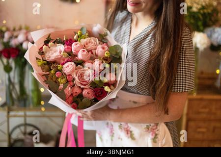 Eine Frau in einem charmanten Blumenladen hält einen atemberaubenden Blumenstrauß aus rosa Rosen und Grün und zeigt das zarte Arrangement mit leuchtenden Farben und nat Stockfoto