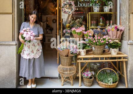 Eine fröhliche Frau steht am Eingang ihres charmanten Blumenladens und hält einen Blumenstrauß in der Hand. Das lebhafte Display zeigt verschiedene Farbfu Stockfoto