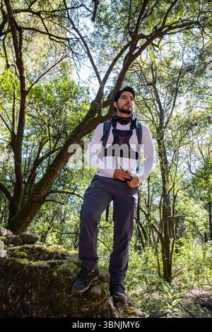 Ein Lateinmann mit Wanderausrüstung steht selbstbewusst auf einem moosbedeckten Felsen, eingebettet in einen üppig grünen Wald. Sonnenlicht filtert durch die Höhe Stockfoto