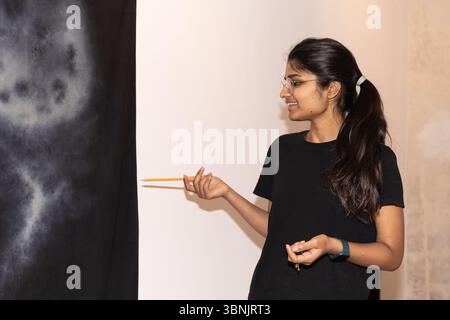 Eine junge indische Künstlerin steht selbstbewusst vor ihrem Kunstwerk, hält einen Bleistift in der Hand, während sie über ihren kreativen Prozess spricht. Sie strahlt Pas aus Stockfoto