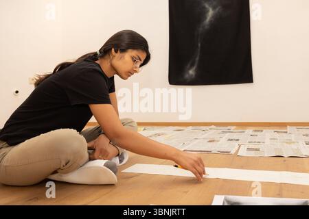 Eine junge indische Künstlerin sitzt im Kreuz auf dem Boden und konzentriert sich auf ihre Arbeit. Umgeben von Papieren und Kunstmaterialien, kreiert sie mit Konzentrat Stockfoto