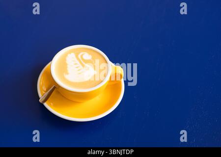 Ein wunderschön gefertigter Latte mit Schwanenmotiv im Schaum, serviert in einer hellgelben Tasse mit Untertasse, auf einer auffälligen blauen Oberfläche, Kreatin Stockfoto