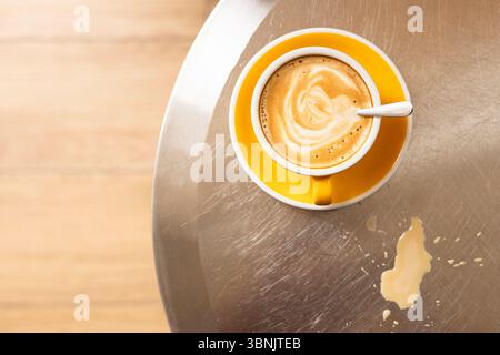 Eine leuchtend gelbe Tasse mit Kaffee sitzt auf einer reflektierenden Metallic-Oberfläche. Daneben trägt ein Kaffeefleck zum rustikalen Charme bei. Die Komposition exu Stockfoto