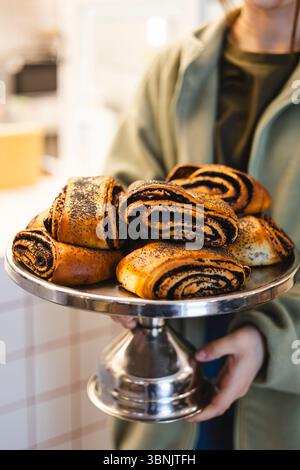 Ein nicht erkennbarer Coffee-Shop-Mitarbeiter, der ein Tablett mit frisch gebackenen Mohnbrötchen präsentiert. Die Umgebung steht für eine gemütliche Bäckerei für kleine Unternehmen Stockfoto