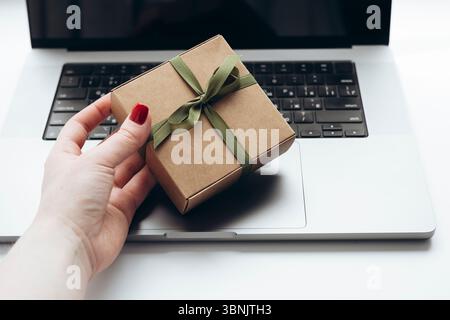 Eine Frau mit gepflegten Nägeln hält eine mit einem grünen Band umwickelte Geschenkbox über einer eleganten Laptop-Tastatur, was einen Online-Geschenkeinkauf oder einen digitalen Gi anregt Stockfoto