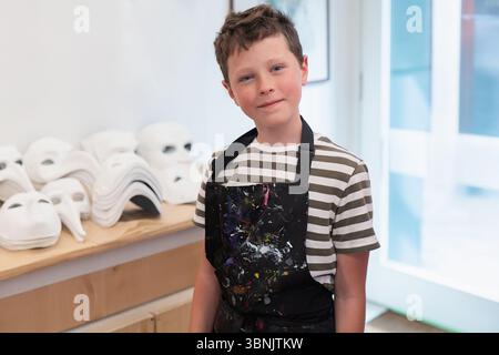 Ein Kind nimmt an einer lebhaften Werkstatt zur Masken-Herstellung in Venedig Teil, trägt eine Schürze mit Farbspritzern und ist umgeben von traditionellen weißen Masken, Expressi Stockfoto