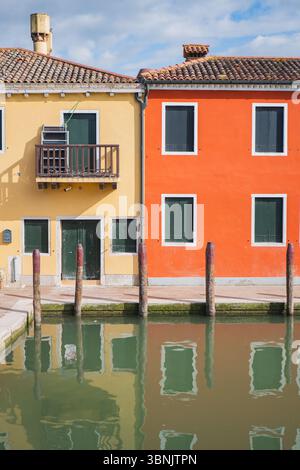 Lebhafte gelbe und orange Häuser spiegeln sich auf einem ruhigen Kanal in der malerischen Stadt Burano und Murano, Venedig. Die sonnige Szene fängt den Charme ein Stockfoto