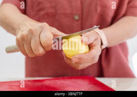 Eine zugeschnittene, detaillierte Ansicht einer älteren Frau, die in einer warmen, heimeligen Küchenatmosphäre eine Kartoffel schält. Ihre erfahrenen Hände arbeiten sorgfältig mit einem Messer, Stockfoto