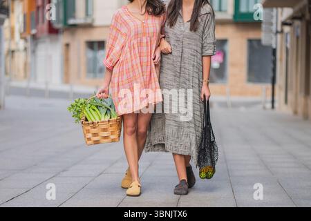 Zwei Freunde genießen einen Sommertag, Arm in Arm auf einer Stadtstraße. Der eine trägt einen Korb mit frischen Erzeugnissen, während der andere einen Netzbeutel enthält. Stockfoto