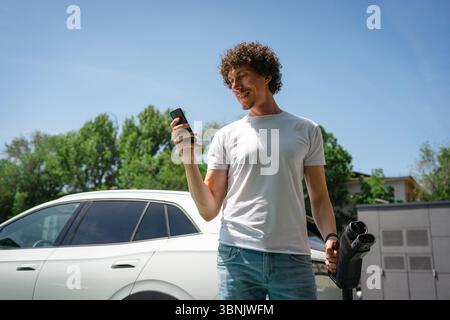 Ein Mann nutzt sein Handy, während er sein Elektroauto in einer Stadt auflädt und stellt eine umweltfreundliche Alternative für den städtischen Nahverkehr unter einem klaren Licht vor Stockfoto