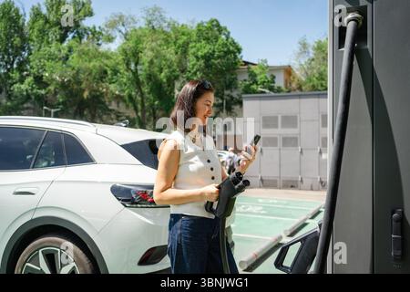 Eine Frau lädt ihr Elektroauto an einer Ladestation in der Stadt auf, während sie ihr Smartphone überprüft, was für modernen und nachhaltigen Stadtverkehr steht Stockfoto