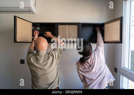 Ein älterer kaukasischer Mann und eine junge Asiatin arbeiten zu Hause an einem DIY-Projekt zusammen, um ein Kabinett zu bauen. Die Teamarbeit und der Fokus heben ihre hervor Stockfoto