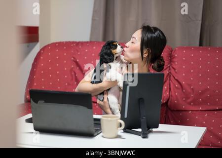 Eine Asiatin arbeitet von zu Hause aus und kümmert sich um ihren verspielten Welpen. Sie verwaltet ihre Aufgaben effizient, indem sie die beruflichen Verantwortlichkeiten ausgleicht Stockfoto