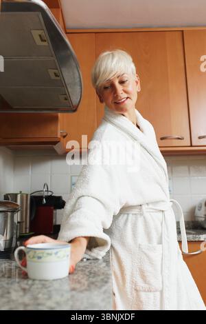 Eine ruhige Morgenszene mit einer Frau in einem weißen Bademantel, die in einer gemütlichen Küche nach einer Kaffeetasse greift und Wärme und Entspannung im Stern zum Ausdruck bringt Stockfoto