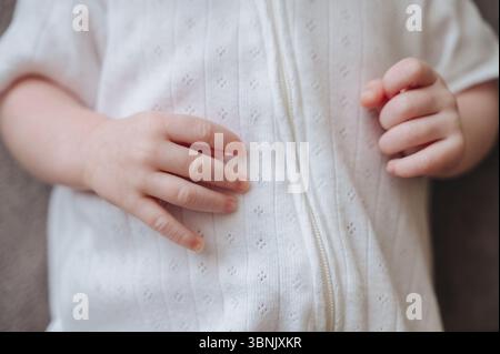 Nahaufnahme der zarten Hände eines einmonatigen Neugeborenen, die sanft auf einem weißen, strukturierten Outfit liegen. Die Babyhände sind weich und friedlich Stockfoto