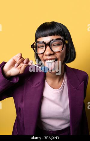 Hispanische junge Frau in lila Jacke, die einen blauen Lutscher vor einem leuchtend gelben Hintergrund genießt. Ihr verspielter Ausdruck und ihre übergroße Brille sorgen für eine besondere Note Stockfoto