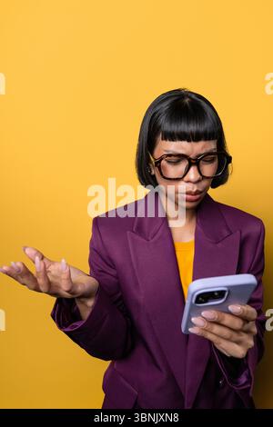 Eine junge hispanische Frau in einer lila Jacke und einer großen Brille sieht verwirrt aus, während sie ihr Smartphone vor einem hellgelben Hintergrund untersucht Stockfoto