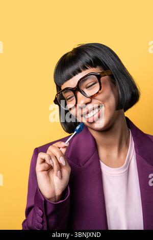 Eine junge hispanische Frau mit kurzen schwarzen Haaren und einer Brille schmeckt einen blauen Lutscher, gekleidet in einem lila Blazer vor einem leuchtend gelben Hintergrund, exsudi Stockfoto