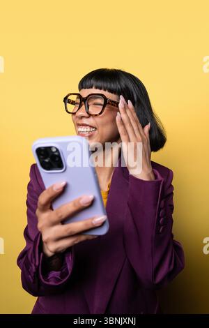 Eine junge hispanische Frau mit kurzen schwarzen Haaren und Brille lacht fröhlich, während sie ein Smartphone hält. Sie trägt einen violetten Blazer gegen eine gelbe Ba Stockfoto