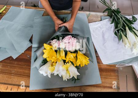 Ein erfahrener Blumenhändler auf Bali arrangiert sorgfältig einen wunderschönen Blumenstrauß aus gelben, weißen und rosa Blumen auf einem Holztisch, der die Kunst der Blumenkunst zeigt Stockfoto