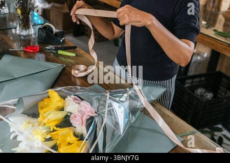 Eine Floristin auf Bali arrangiert geschickt einen lebendigen Blumenstrauß mit gelben Lilien und rosa Rosen auf einem Holztisch und zeigt ihre Kunstfertigkeit Stockfoto