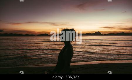 Seitenansicht der Silhouette einer nicht erkennbaren Frau, die bei Sonnenuntergang an einem ruhigen philippinischen Strand steht, mit Inseln im Hintergrund. Stockfoto
