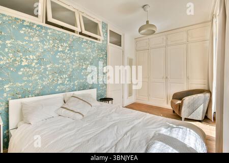 Schlafzimmer mit Sonnenlicht, eleganter geblümter Tapete, weißer Bettwäsche und moderner Einrichtung. Mit eingebauten Schränken und einem bequemen Sessel für eine ruhige Atmosphäre Stockfoto